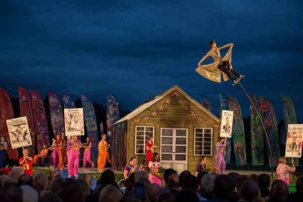 The Heritage Flame Ceremony in Stoke Mandeville. A shed-like building with bunting stands in front of colourful flags on an open-air stage. Performers with signs depicting pictures and names of paralympic athletes stand alongside others both standing and in wheelchairs, holding flowers. On a bendy fibreglass pole, a performer (Amelia Cavallo) sways holding a shiny gold coat out wide, high above the roof of the shed. At the bottom of this page is a video of this performance with audio commentary.